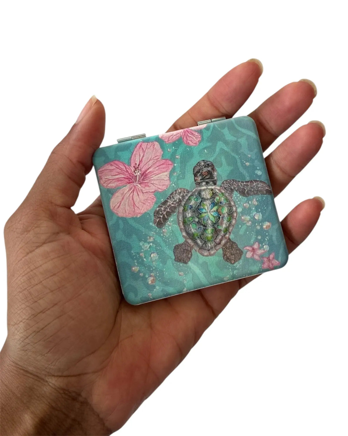 Miroir de poche fermé tenu dans une main, décoré d’une tortue marine et de fleurs roses