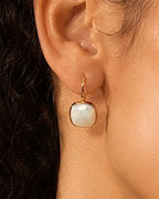 Boucle d'oreille dorée à cabochon carré en nacre blanche portée par une femme aux cheveux bouclés