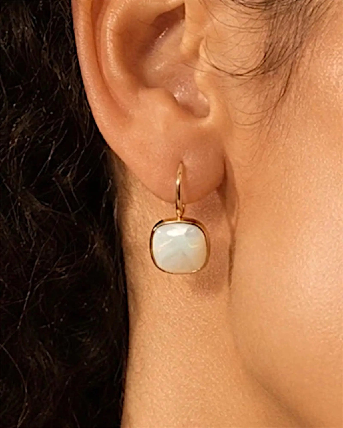 Boucle d'oreille dorée à cabochon carré en nacre blanche portée par une femme aux cheveux bouclés