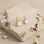 Boucle d'oreille dorée à cabochon carré en nacre blanche portée par une femme aux cheveux bouclés