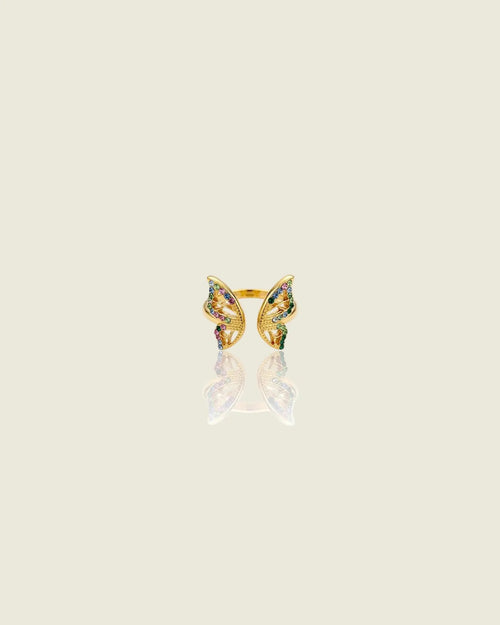 Bague ouverte dorée en forme de papillon, ornée de pierres multicolores, sur fond beige avec reflet.