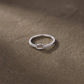 Bague fine argentée ornée de strass et d’un motif carré ajouré, posée sur un fond brun texturé.