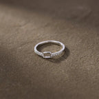 Bague fine argentée ornée de strass et d’un motif carré ajouré, posée sur un fond brun texturé.