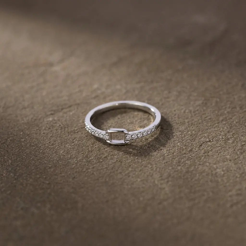 Bague fine argentée ornée de strass et d’un motif carré ajouré, posée sur un fond brun texturé.