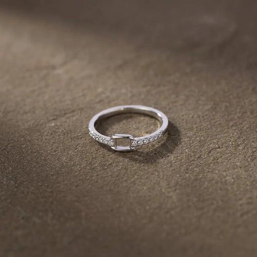 Bague fine argentée ornée de strass et d’un motif carré ajouré, posée sur un fond brun texturé.