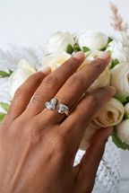 Bague ouverte en acier inoxydable argenté ornée de deux cœurs, portée au majeur devant un bouquet de roses blanches