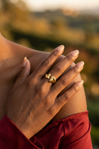 Bague ouverte dorée ornée de deux cœurs, portée au majeur sur une main posée sur l’épaule, en lumière naturelle