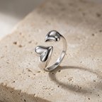 Bague ouverte duo de cœurs en acier inoxydable argenté, posée sur une pierre beige avec ses deux cœurs bombés visibles.