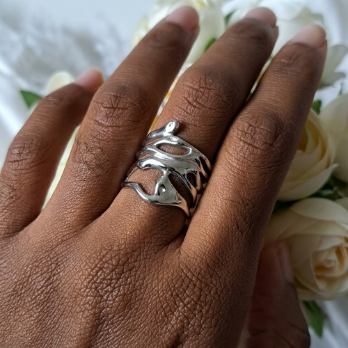 Bague large argentée en acier inoxydable aux lignes ondulées et ajourées, portée au majeur devant des fleurs blanches